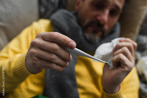 selective focus of electronic thermometer in hand on sick man suffering from fever on blurred background.