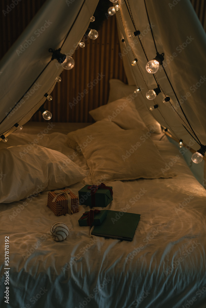 Gifts on camping tent bed decorated with Christmas lights. Kid's bedroom. Winter holidays. Evening atmosphere.