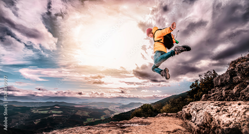 Obraz premium Hiker with backpack jumping on top of a mountain rejoices at having achieved the goal - The successful man at the end of a day of hiking in the mountains reaches the summit enjoying the victory