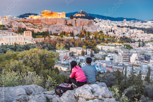 Obraz na płótnie Middle aged couple of travellers from Asia sitting at stone admiring fabulous sunset over ancient Athen's city downtown, view at Parthenon and Agora hill, during twilights. Athens, Greece, Europe. w salonie