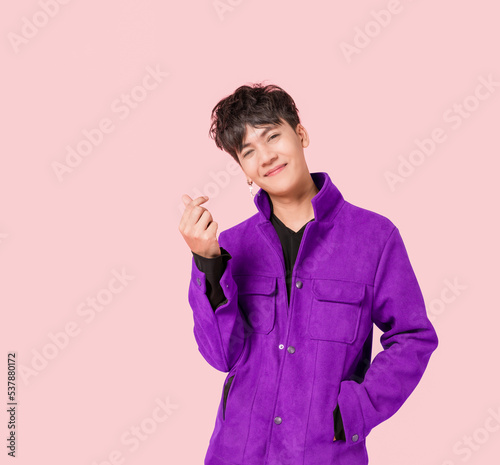 Portrait of a happy Asian handsome man in fashionable purple jacket clothing standing smiling using fingers to make a mini heart sign isolated on pink background.