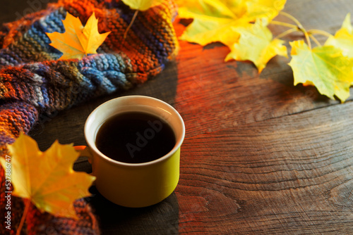 A yellow mug of hot coffee on a wooden table with yellow autumn leaves and a warm knitted scarf. copy space