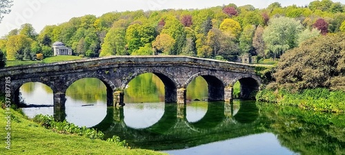 Park with an old stone bridge over a river with beautiful trees in the background © Michael Fining/Wirestock Creators