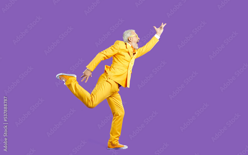 Funny senior man dancing, singing and having fun in studio. Side view of happy energetic bearded mature man wearing bright yellow party suit dancing isolated on solid purple colour background