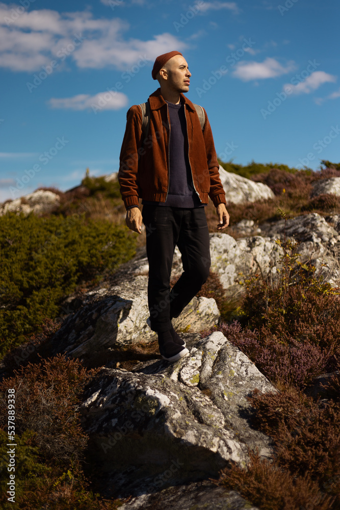 A caucasian man wearing a beanie and a brown jacket with a backpack walking on rocks outdoors.