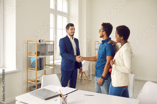 Happy ethnic couple shake hand get acquainted with architect or designer in office. Excited clients or buyers handshake realtor or broker close deal or agreement at meeting. House buy or rent.