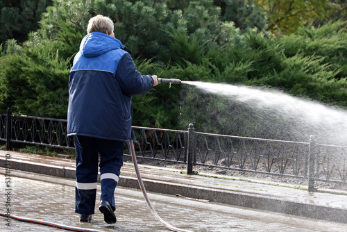 Woman worker in blue uniform watering the sidewalk with a hose. Street cleaning and disinfection in autumn city park