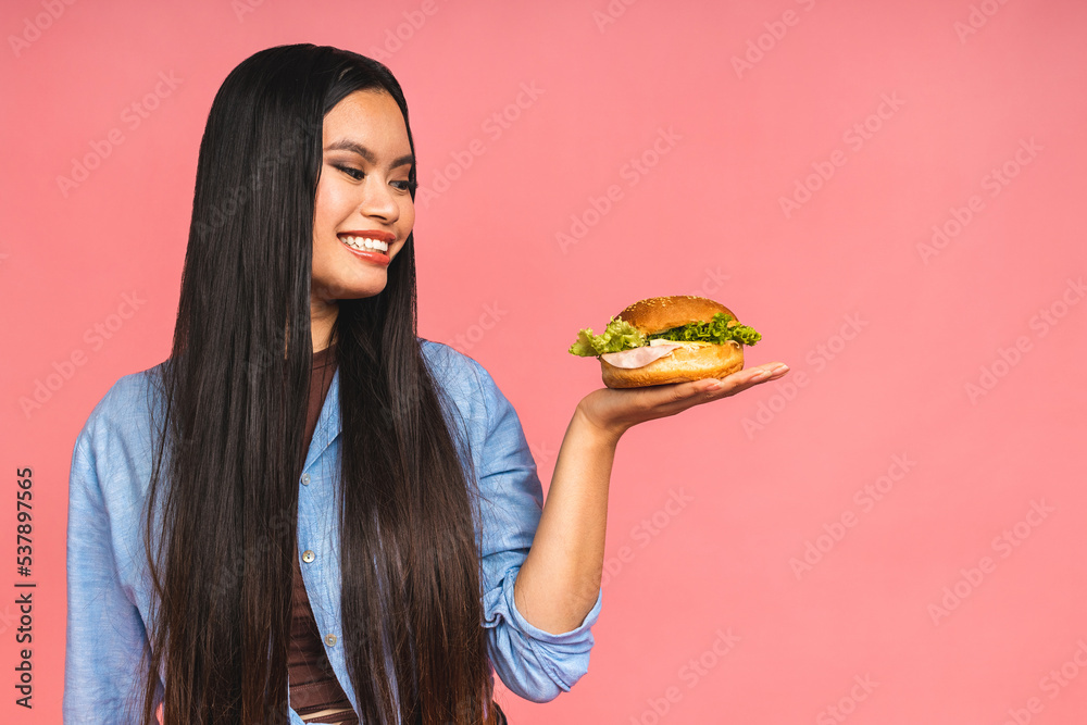 Young beautiful asian japanese chinese woman eating sandwich or big burger with satisfaction. Girl enjoys tasty hamburger takeaway, diet concept, standing isolated over pink background. Young beautiful asian japanese chinese woman eating sandwich or big burger with satisfaction. Girl enjoys tasty hamburger takeaway, diet concept, standing isolated over pink background.