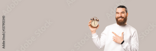 Panorama shot of a bearded man pointing at the alarm clock he is holding. photo