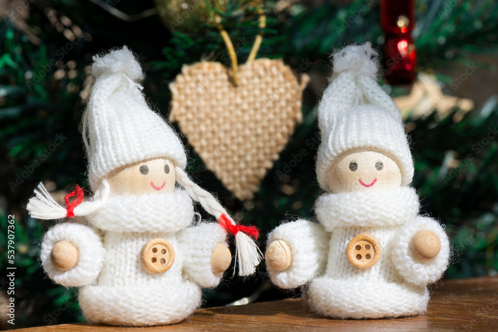 Christmas composition with two wooden dolls boy and girl in white knitted New Year's themed clothes and hats. Blurred сhristmas toy in the shape of a heart on a Christmas tree. Selective focus.