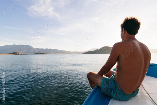 Man in swimming shorts sitting on boat with legs overboard. photo