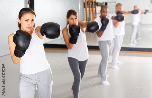 Portrait of active hispanic woman in boxing gloves mastering self defense techniques in group, practicing punches in training room © JackF