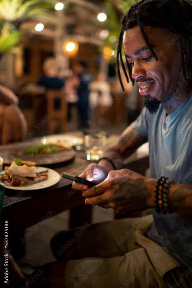 Young black man with tattoos using phone at dinner