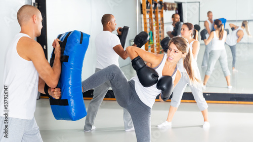 Young focused woman wearing boxing gloves working with trainer during self-defense course  practicing foot strikes on kick shield
