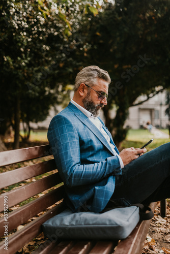 Modern senior businessman using smartphone for online meeting while sitting on bench