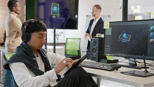 Wallpaper Mural Asian male artist works in headphones in a studio for the development of computer games and applications. In the background, a team of developers are discussing a 3D model. Torontodigital.ca