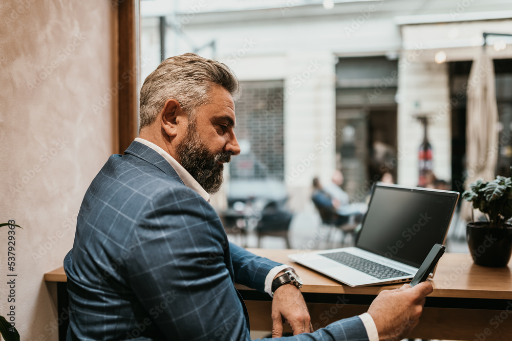 Modern senior businessman sitting in cafe and using laptop and smartphone