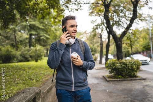 One young adult caucasian man walking in the city town near park using mobile phone smartphone talk with cup of coffee in autumn or spring day happy male tourist standing alone real people copy space