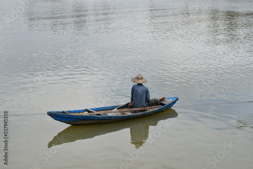 A Vietnamese fisherman in a round Vietnamese hat is fishing in a long fishing boat.