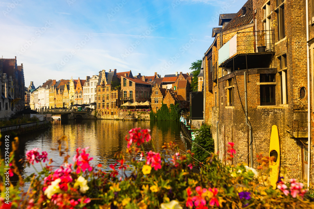 Scenic cityscape of Ghent with traditional Flemish style brick townhouses decorated with colorful blooming flowers on banks of Leie river on sunny summer day, Belgium.
