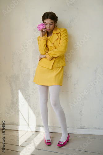 Young girl in a yellow suit with a pink flower near a white wall.