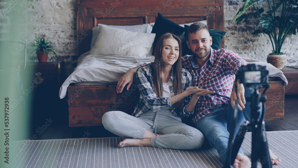 Naklejka premium Adorable young couple is recording video for social media with camera, they are talking to followers sitting on floor of loft style bedroom wearing casual clothes.