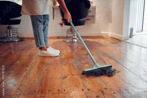 Hairdresser woman, sweeping hair and cleaning salon or barbershop wooden floor after cutting a hairstyle or doing a trim. Feet of female worker with broom for dirt, dust and mess for clean workplace