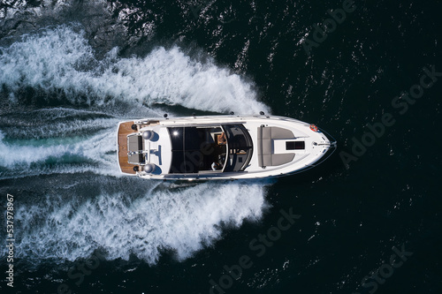 Luxurious large yacht with people moving quickly on dark blue water making a white trail behind the boat. Aerial view of the sea and the boat. Beautiful natural seascape in summer. © Berg