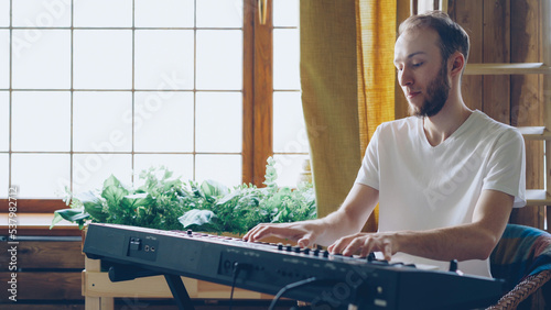 Cheerful young man amateur musician in casual clothes is playing keyboard at home. Musical instruments, attractive people, hobby and creativity concept.
