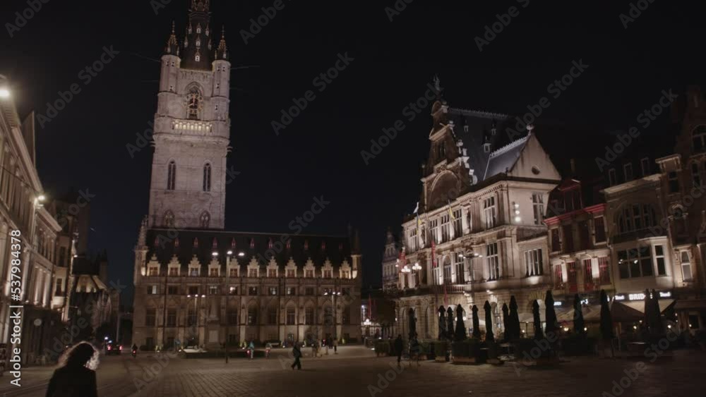 Panning past beautiful square to Belfort (Belfry) tower in Gent, Belgium at night. People are walking around the busy Sint-Baafsplein which is surrounded by monumental and historical buildings Panning past beautiful square to Belfort (Belfry) tower in Gent, Belgium at night. People are walking around the busy Sint-Baafsplein which is surrounded by monumental and historical buildings
