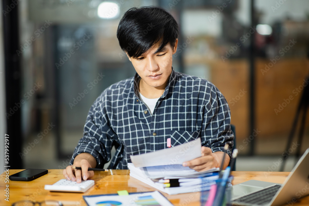 A young financial market analyst works in the office with his laptop while sitting at a wooden table. businessman analyzing graph document in hand. A young financial market analyst works in the office with his laptop while sitting at a wooden table. businessman analyzing graph document in hand.