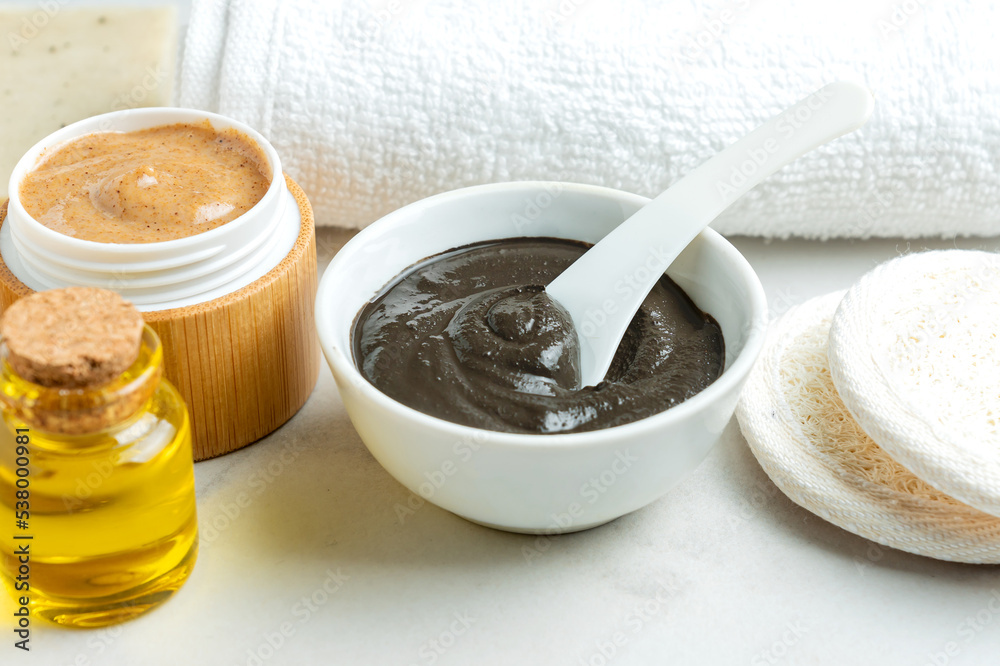 Homemade clay mask for the face and body in a white bowl nect to scrub and oil in bottles. Reusable lufa pads for mask removing and cotton towel on a table. Spa procedures at home. Healthy leaving