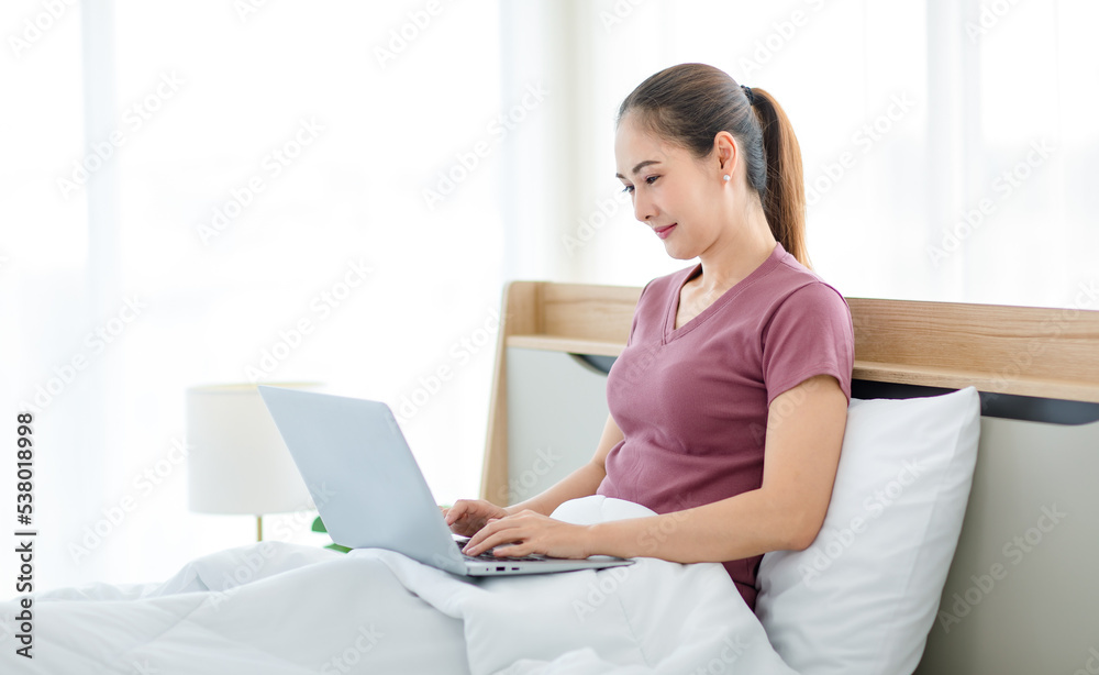 Millennial Asian young happy cheerful female housewife sitting smiling on bed and pillow under thick warm blanket using laptop notebook computer browsing surfing internet shopping online in bedroom Millennial Asian young happy cheerful female housewife sitting smiling on bed and pillow under thick warm blanket using laptop notebook computer browsing surfing internet shopping online in bedroom