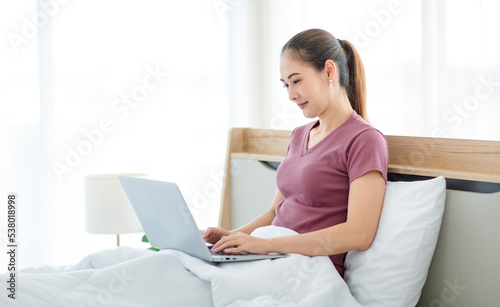 Millennial Asian young happy cheerful female housewife sitting smiling on bed and pillow under thick warm blanket using laptop notebook computer browsing surfing internet shopping online in bedroom