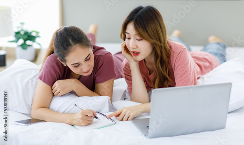 Millennial Asian young happy cheerful female teenager LGBTQ lesbian lover couple laying down together on bed and pillow learning studying online via laptop computer and writing short note on notebook