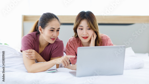Millennial Asian young happy cheerful female teenager LGBTQ lesbian lover couple laying down together on bed and pillow learning studying online via laptop computer and writing short note on notebook