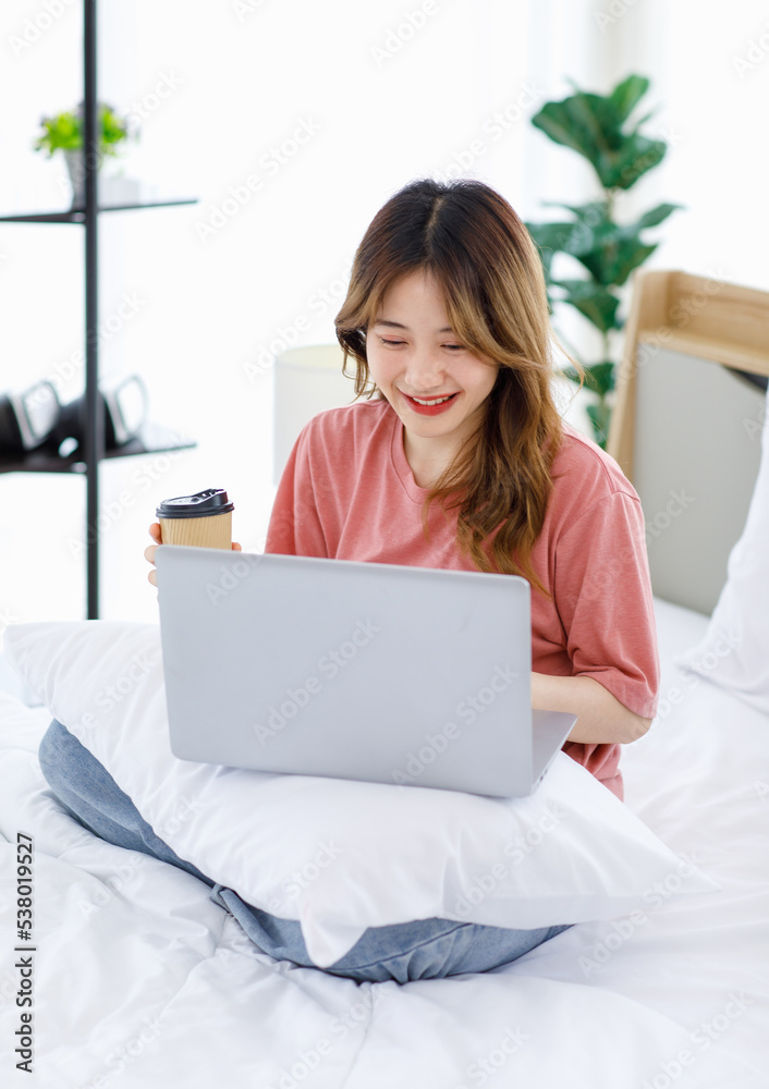 Millennial Asian young happy cheerful female teenager sitting smiling on cozy bed in bedroom at home holding touchscreen tablet computer video call with friend while drinking hot coffee in morning