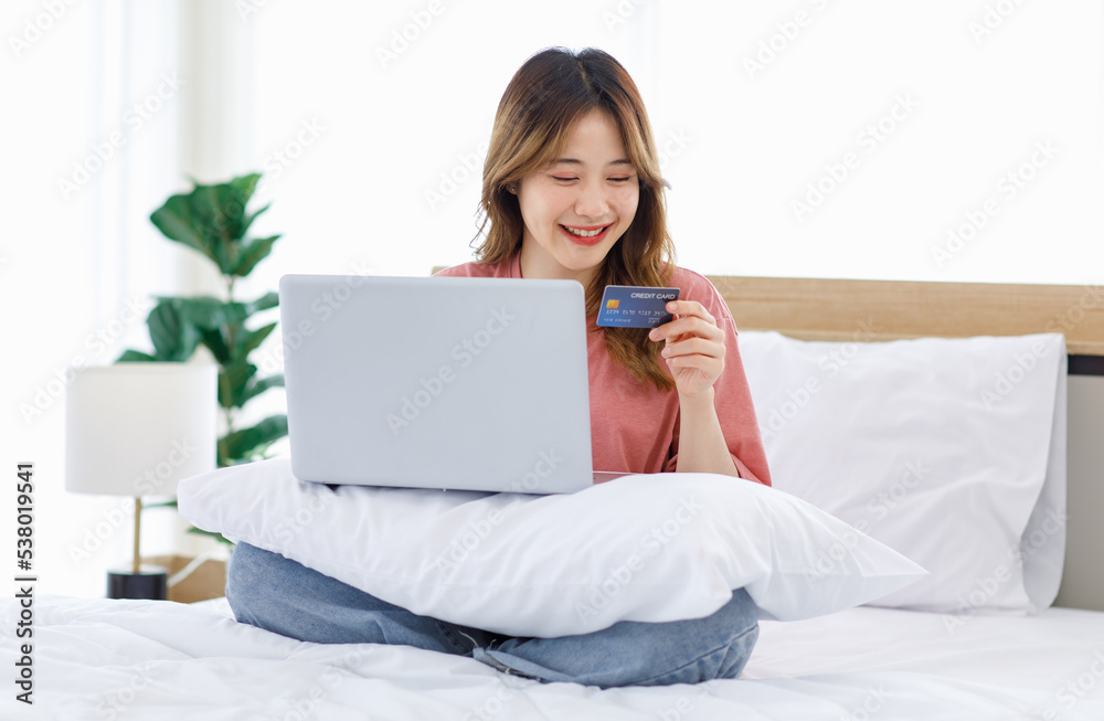 Millennial Asian young satisfied excited female teenager laying smiling under blanket on bed in bedroom at home in morning use credit card making payment shopping online via laptop notebook computer Millennial Asian young satisfied excited female teenager laying smiling under blanket on bed in bedroom at home in morning use credit card making payment shopping online via laptop notebook computer