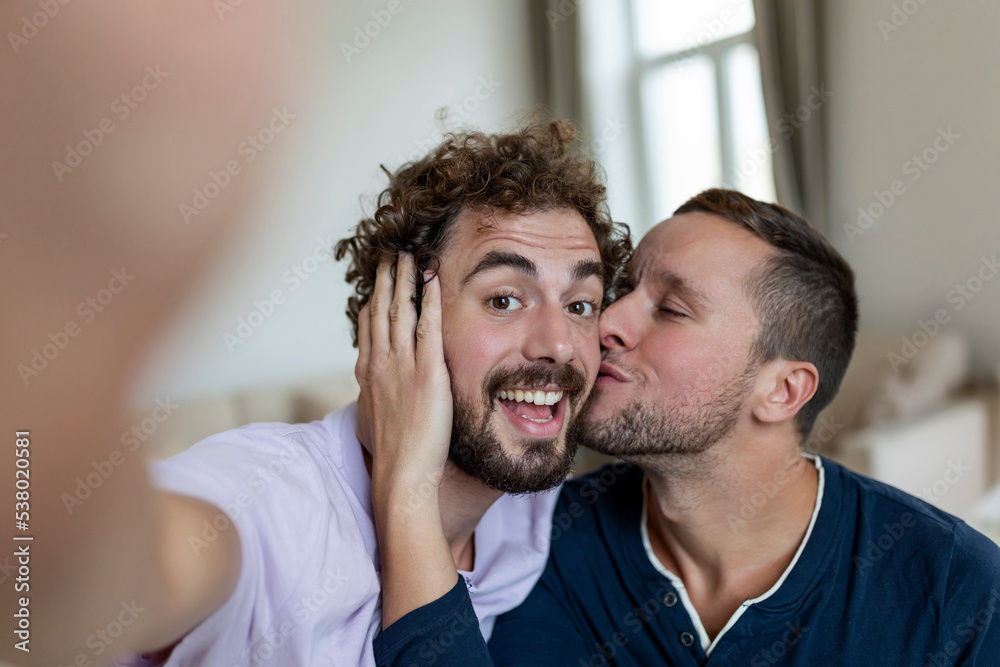 Obraz premium Handsome gay man sitting together at home taking selfie with mobile phone. Gay man with his partner sitting by taking a self portrait using smart phone.