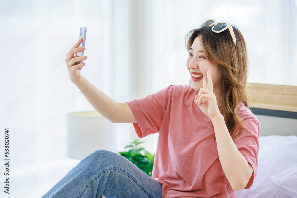 Millennial Asian young happy cheerful female teenager with sunglasses sitting smiling on cozy bed in bedroom at home holding smartphone taking selfie photo while packing clothes for holiday vacation Millennial Asian young happy cheerful female teenager with sunglasses sitting smiling on cozy bed in bedroom at home holding smartphone taking selfie photo while packing clothes for holiday vacation