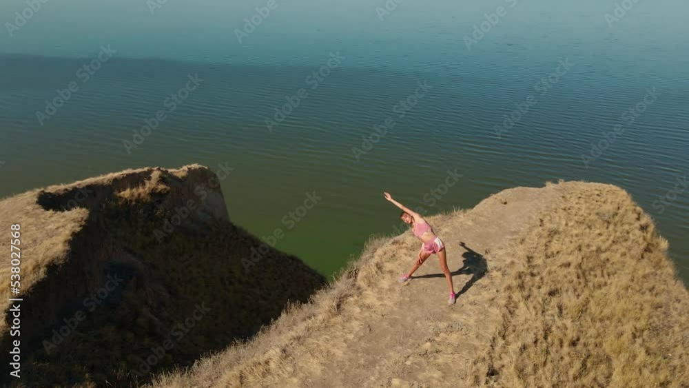 aerial view of stretching exercises outdoors. drone flight around fitness woman jogger doing exercises and stretches at morning on edge of clay ridge canyon on sea shore aerial view of stretching exercises outdoors. drone flight around fitness woman jogger doing exercises and stretches at morning on edge of clay ridge canyon on sea shore