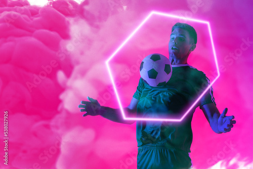 Caucasian male player playing with soccer ball over illuminated hexagon against pink neon smoke