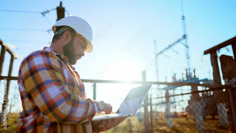 Builder stands in protective helmet in field against background of high telecommunications tower and uses digital tablet to monitor and develop logistics plan building this territory. Businessman. Builder stands in protective helmet in field against background of high telecommunications tower and uses digital tablet to monitor and develop logistics plan building this territory. Businessman.