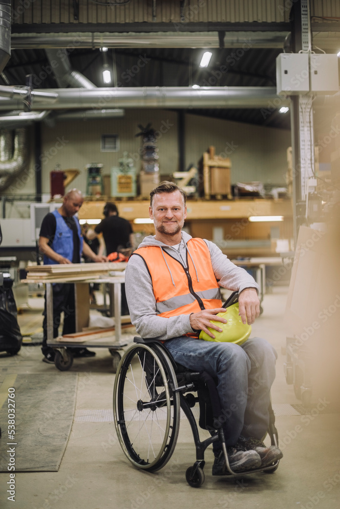Portrait of smiling carpenter sitting on wheelchair with colleagues working in background at warehouse