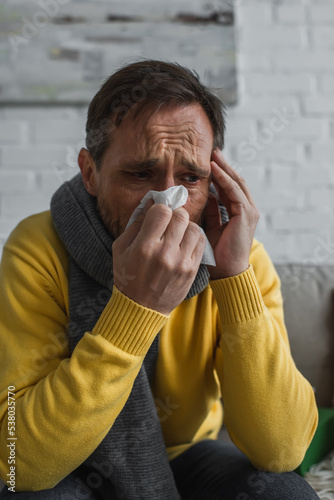 ill man in warm scarf sneezing in paper napkin and suffering from migraine at home.