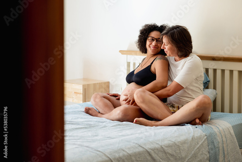 Woman holding and touching her pregnant girlfriend belly while sitting on the bed together at home. Pregnancy and motherhood concept.
