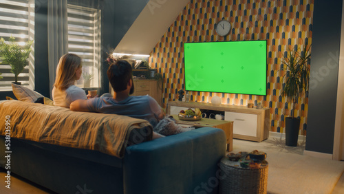 Couple sitting on sofa in living room, talking, watching breaking news, movie or series on TV. Wife and husband resting and chilling at home in the evening together. Green screen. Chromakey.