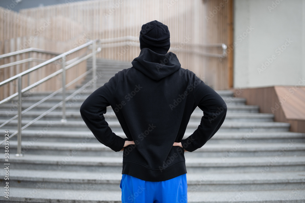 A man in a winter running uniform stands in front of the steps up. Rear view of hooded sportsman before run upstairs. A man in a winter running uniform stands in front of the steps up. Rear view of hooded sportsman before run upstairs.