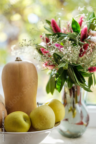 Close up shot of an autumn composition, bouquet of flowers in a vase, butternut squashes, apples in a bowl.
