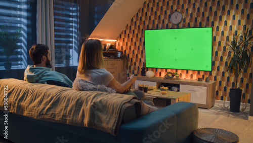 Woman switching channels with remote control. Couple sitting on couch in living room, talking, watching breaking news, movie or series on TV. Resting at home in the evening. Green screen. Chromakey.